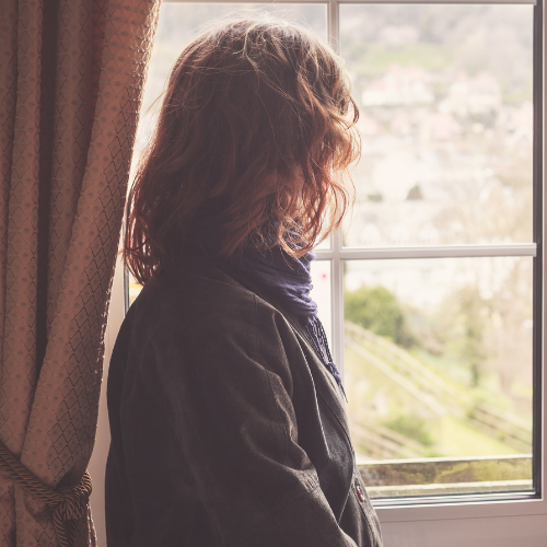 A person with curly hair standing near a window, looking outside.