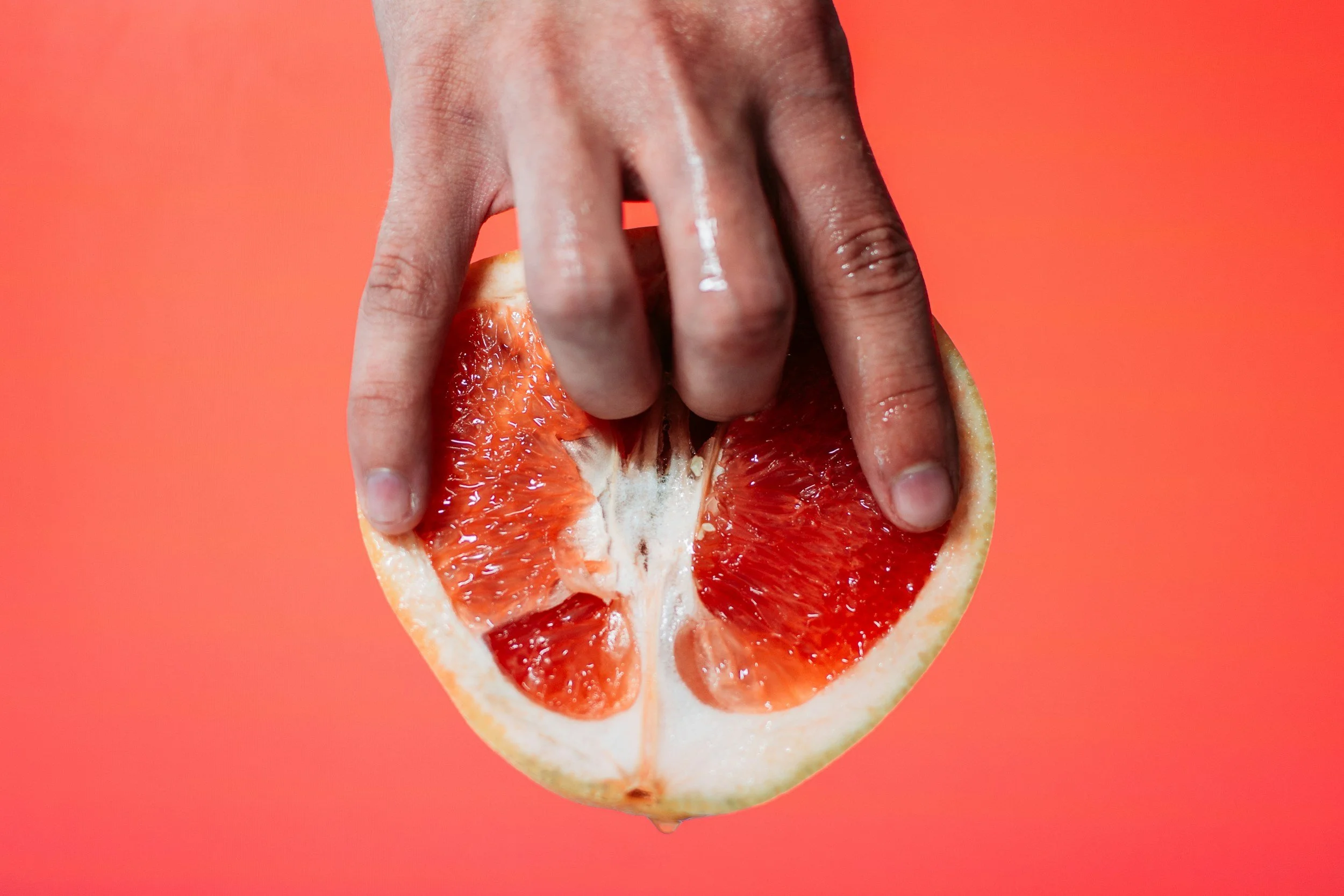 A hand holding a halved red grapefruit against a coral pink background.
