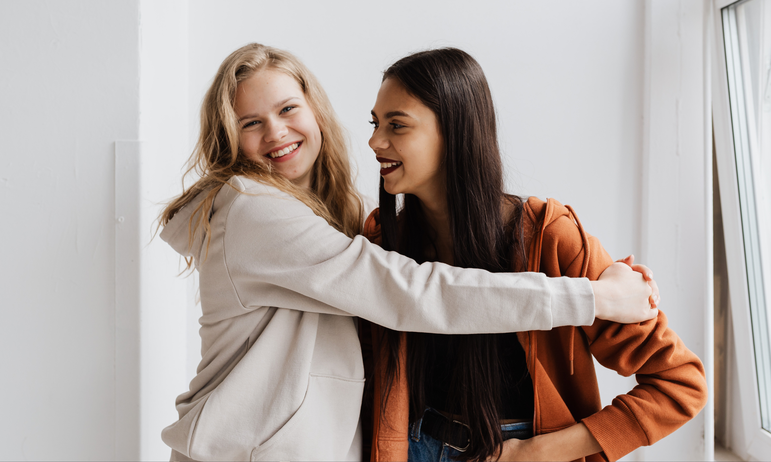 Two young women hugging and smiling at each other indoors near a window.