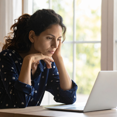 Woman with curly hair wearing a dark blue shirt with white polka dots, sitting at a table and working on a laptop near a window.