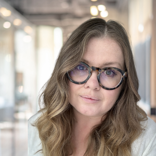 Close-up of a woman with long wavy brown hair and glasses, smiling softly in a bright indoor space.