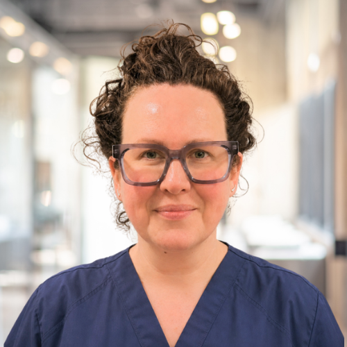 A woman with dark curly hair and glasses wearing a navy blue medical scrub top, standing in a well-lit medical or laboratory setting.