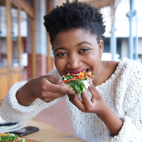 A woman with short curly hair eating a slice of pizza with vegetables at a restaurant.