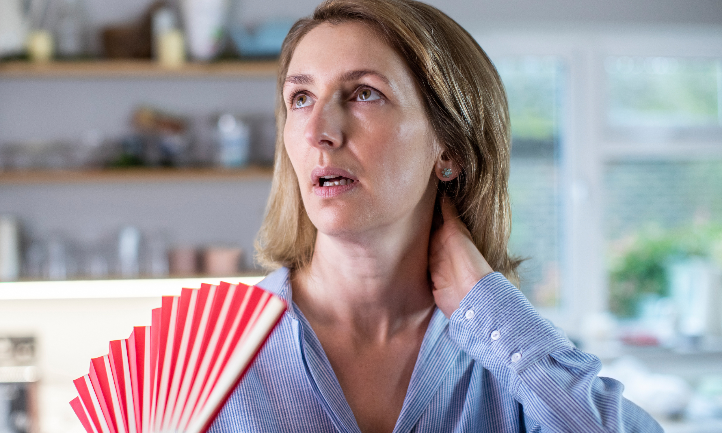 A woman with light brown hair and blue eyes holding a red paper fan, wearing a light blue striped shirt, standing in a kitchen with blurred shelves and a window in the background.