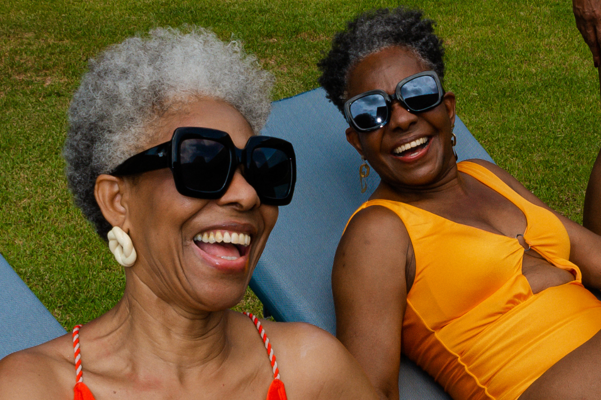 Two smiling women, one with gray curly hair and large sunglasses, the other with short black hair wearing sunglasses, both lying on blue lounge chairs on the grass during a sunny day.