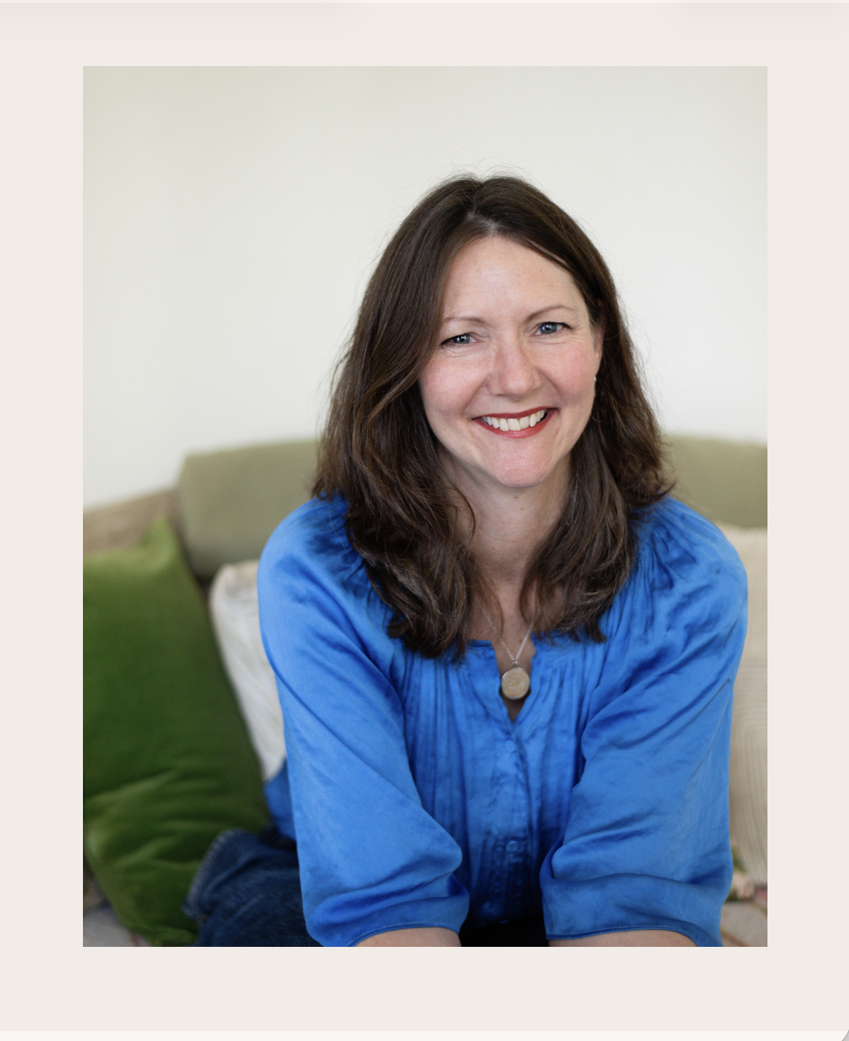 A caring holistic female doctor with brown curly hair wearing a blue blouse and a circular pendant necklace, smiling and sitting on a couch with green and beige pillows in the background.