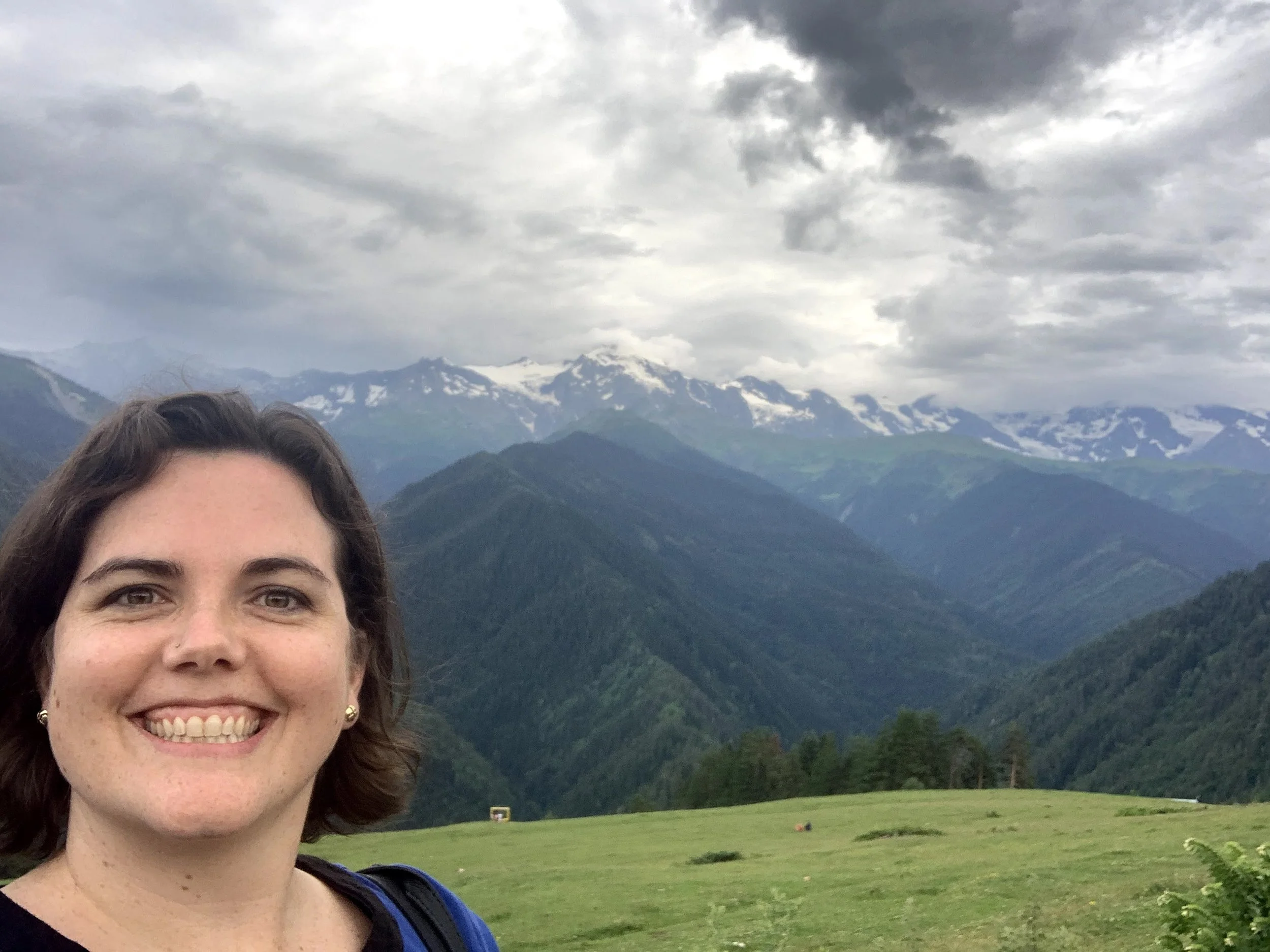 A woman smiling in the foreground with lush green mountains and snowy peaks under a cloudy sky in the background.