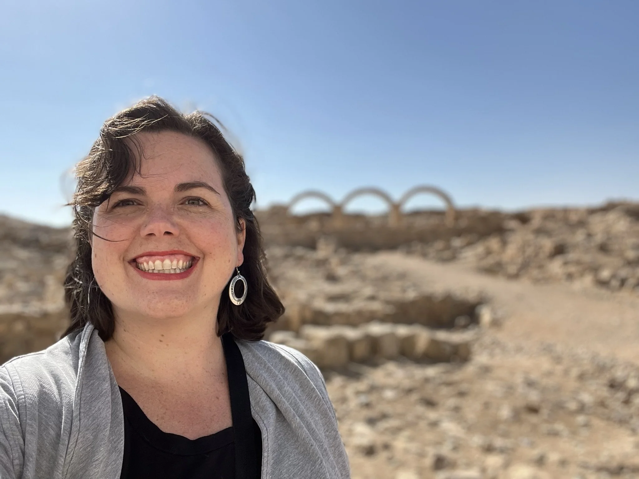A smiling woman with dark hair and earrings taking a selfie in a desert landscape with an ancient stone arch bridge in the background under a clear blue sky.