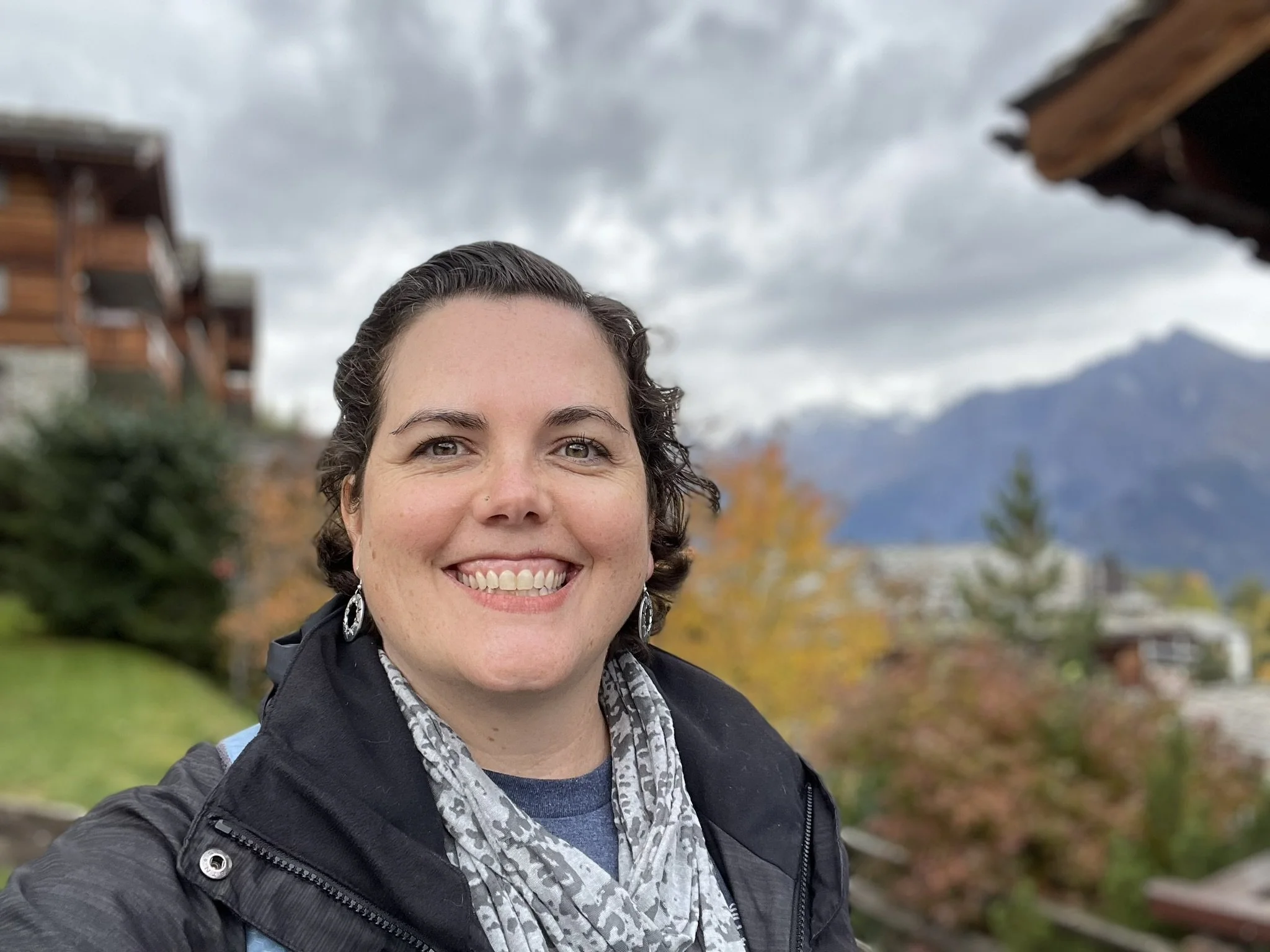 A woman taking a selfie outdoors with mountains, trees with autumn leaves, and houses in the background under cloudy skies.
