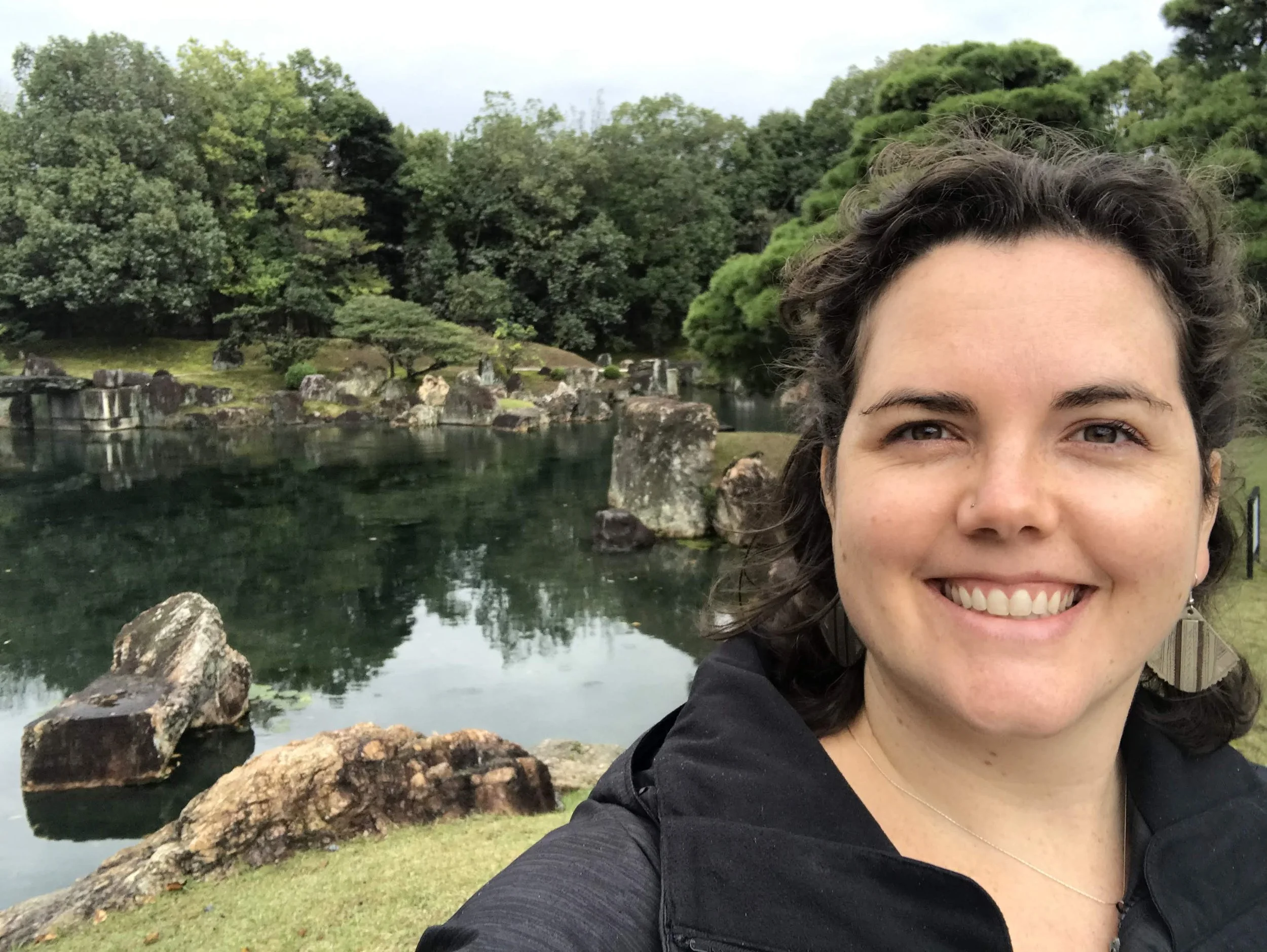 A woman smiling, taking a selfie in front of a scenic Japanese-style garden with a pond, rocks, and lush green trees.
