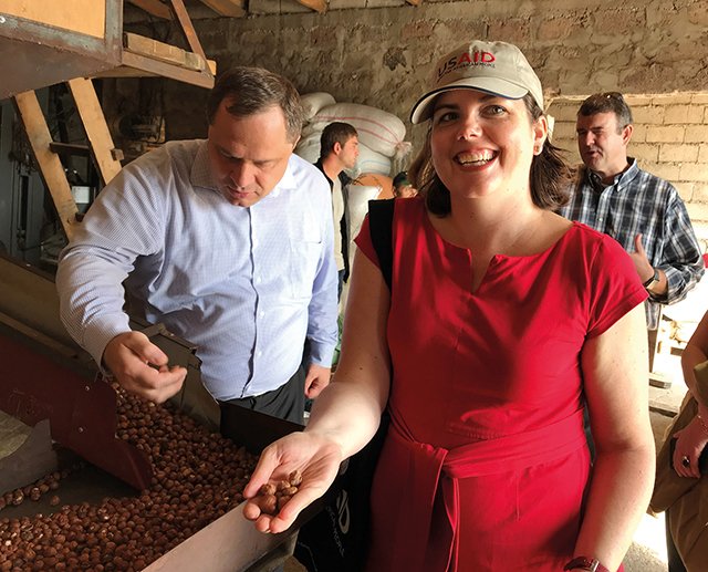 A smiling woman in a red dress and USAID cap holding coffee beans at a coffee processing facility, with other people and sacks of coffee in the background.
