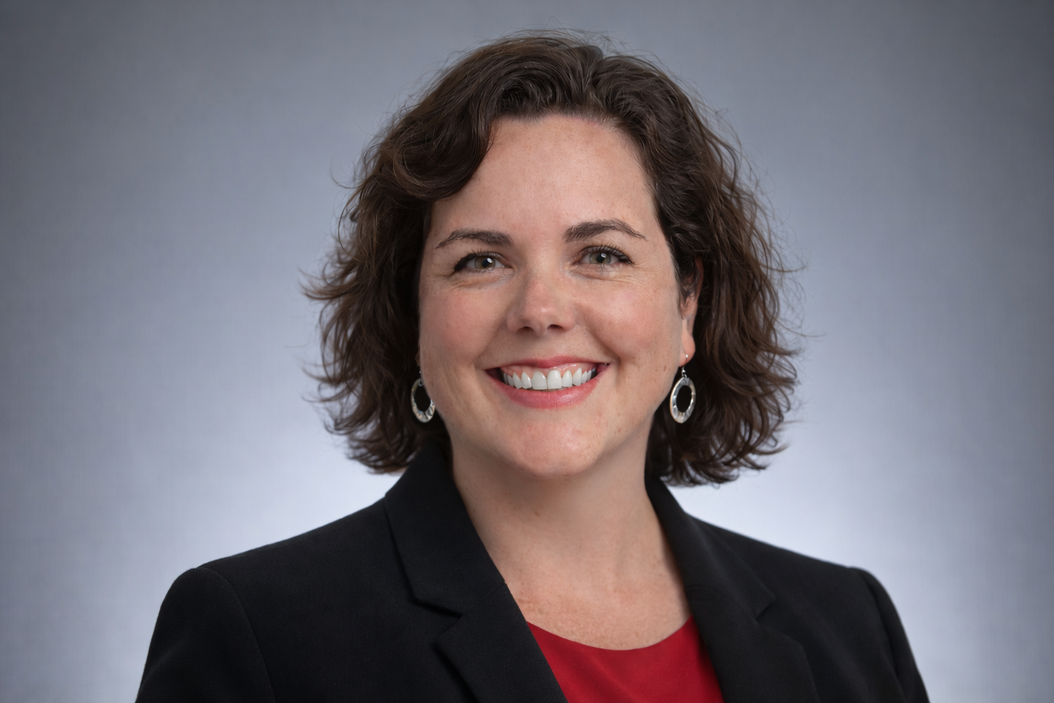 Marika Olson - A woman with short, curly brown hair smiling at the camera, dressed in a black blazer and red top, wearing silver earrings, against a gray background.