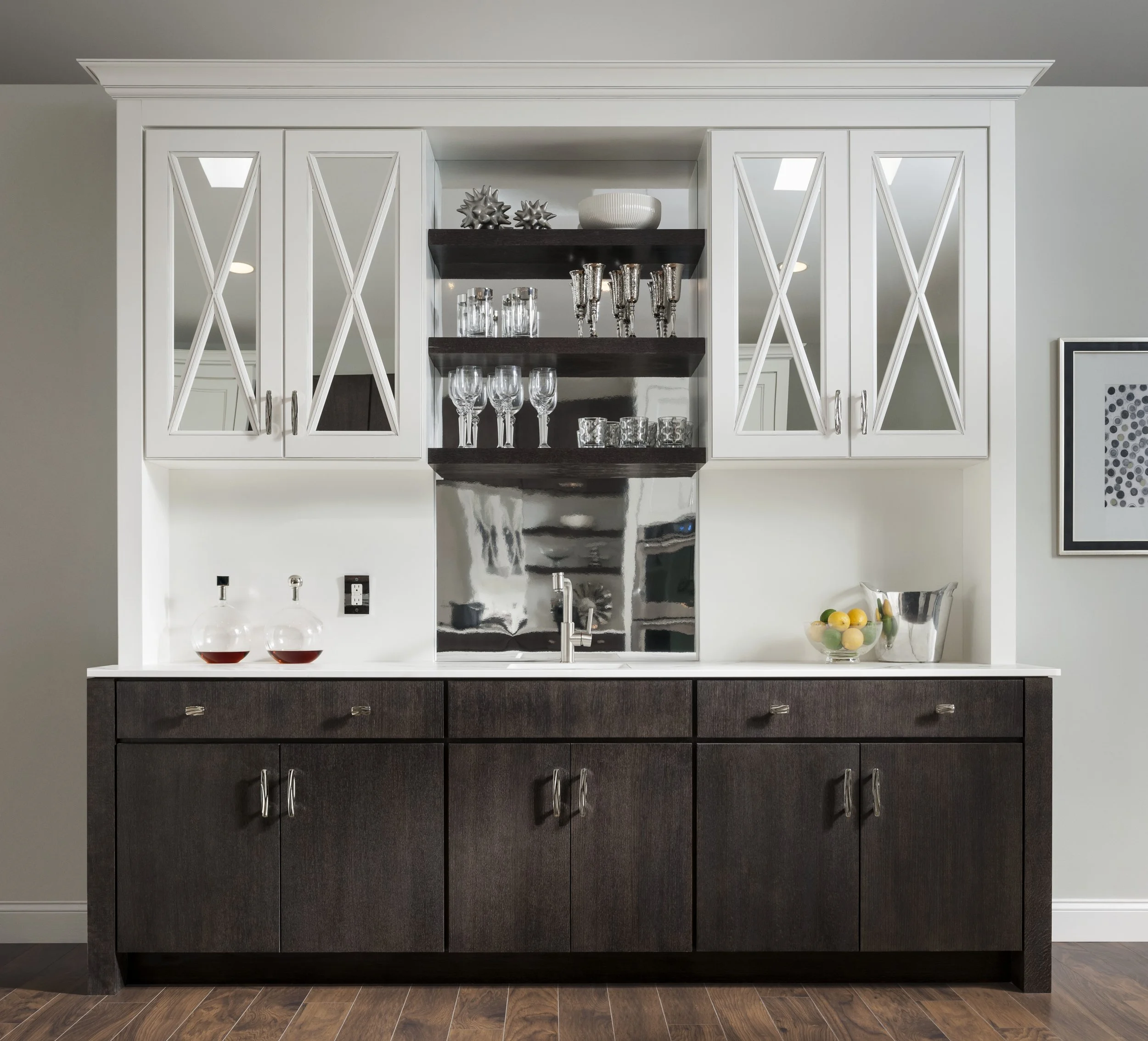 A modern bar cabinet with white upper cabinets with glass-paneled doors, open dark wood shelves in the middle holding glassware, a dark wood lower cabinet, and a white countertop with decorative bottles, a bowl of lemons, and a metal ice bucket.