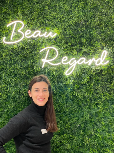 Une femme opticienne souriante avec un pull noir, debout devant un mur recouvert de végétation verte, avec un panneau lumineux indiquant 'Beau Regard' en cursive éclairée.