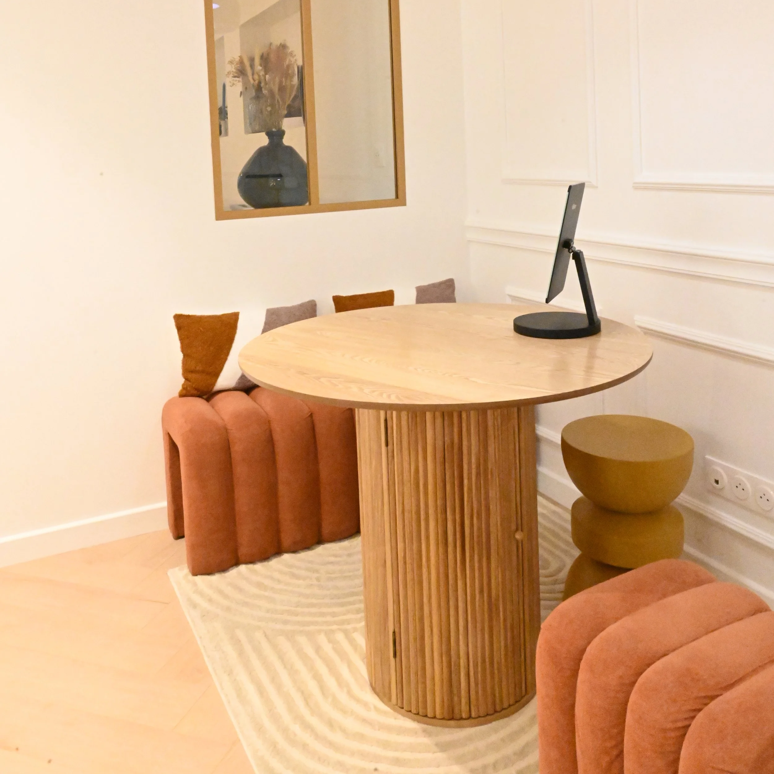 Un coin salon avec un canapé en velours orange, un tapis beige, un mur blanc avec moulures, et une table ronde en bois avec un support vertical en bois. Sur la table, un écran d'ordinateur. Des coussins de différentes couleurs sont posé sur le canapé.