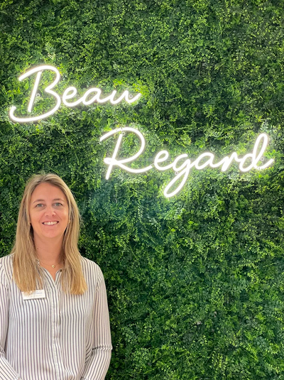 Une femme opticienne souriante se tient devant un mur de végétation avec un neon indiquant 'Beau Regard'.