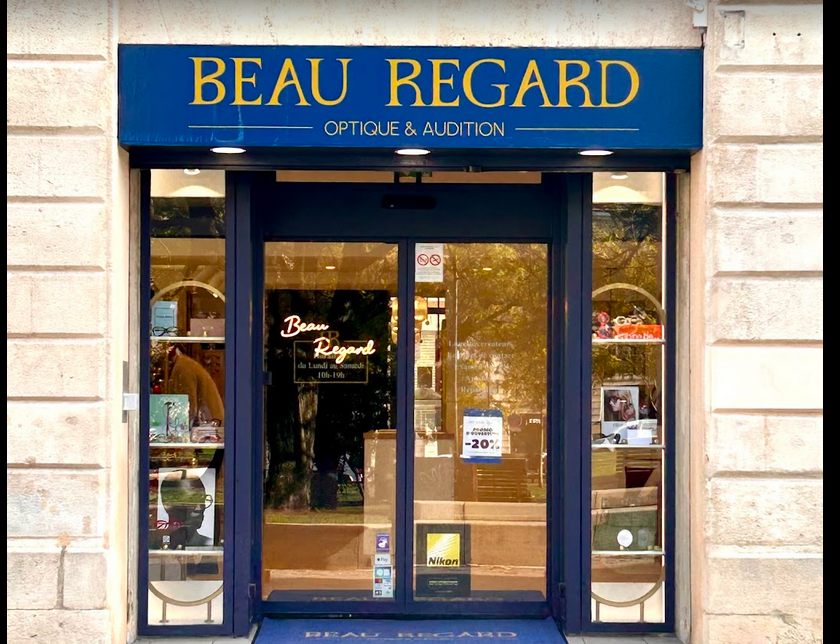 Façade d'un magasin optique nommé BEAU REGARD, avec une enseigne bleue avec lettres jaunes, vitres sur les côtés affichant des produits, et une porte en verre.