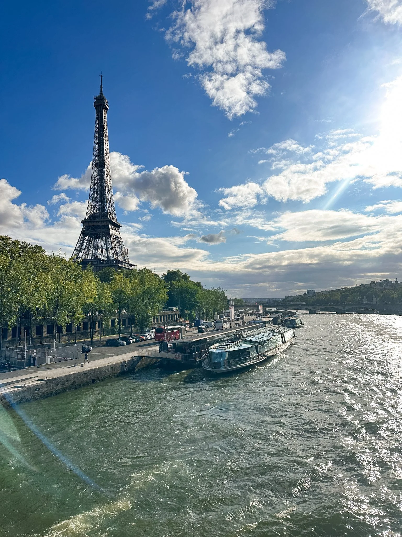 View of the Eiffel Tower along the Seine River, with a boat docked at the riverbank and partly cloudy sky.