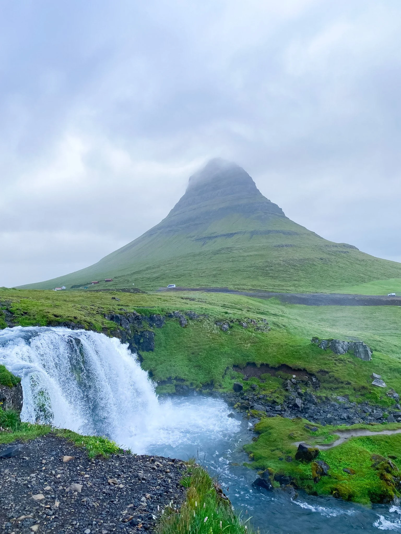 Scenic view of a waterfall flowing into a river with green grass and moss-covered rocks, a mountain with a cloud-covered peak in the background, and a few cars and small houses on the road.