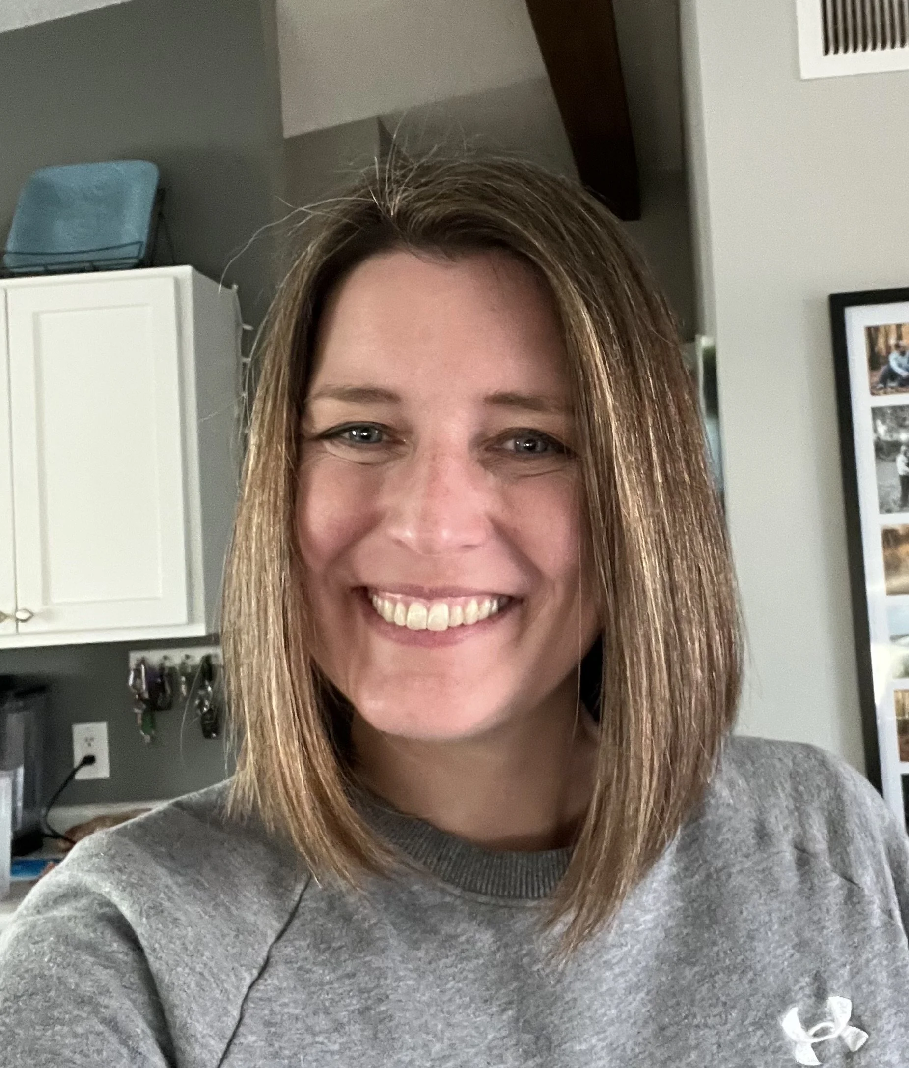 A man in a black blazer and checkered shirt smiling outdoors with blurred greenery in the background.