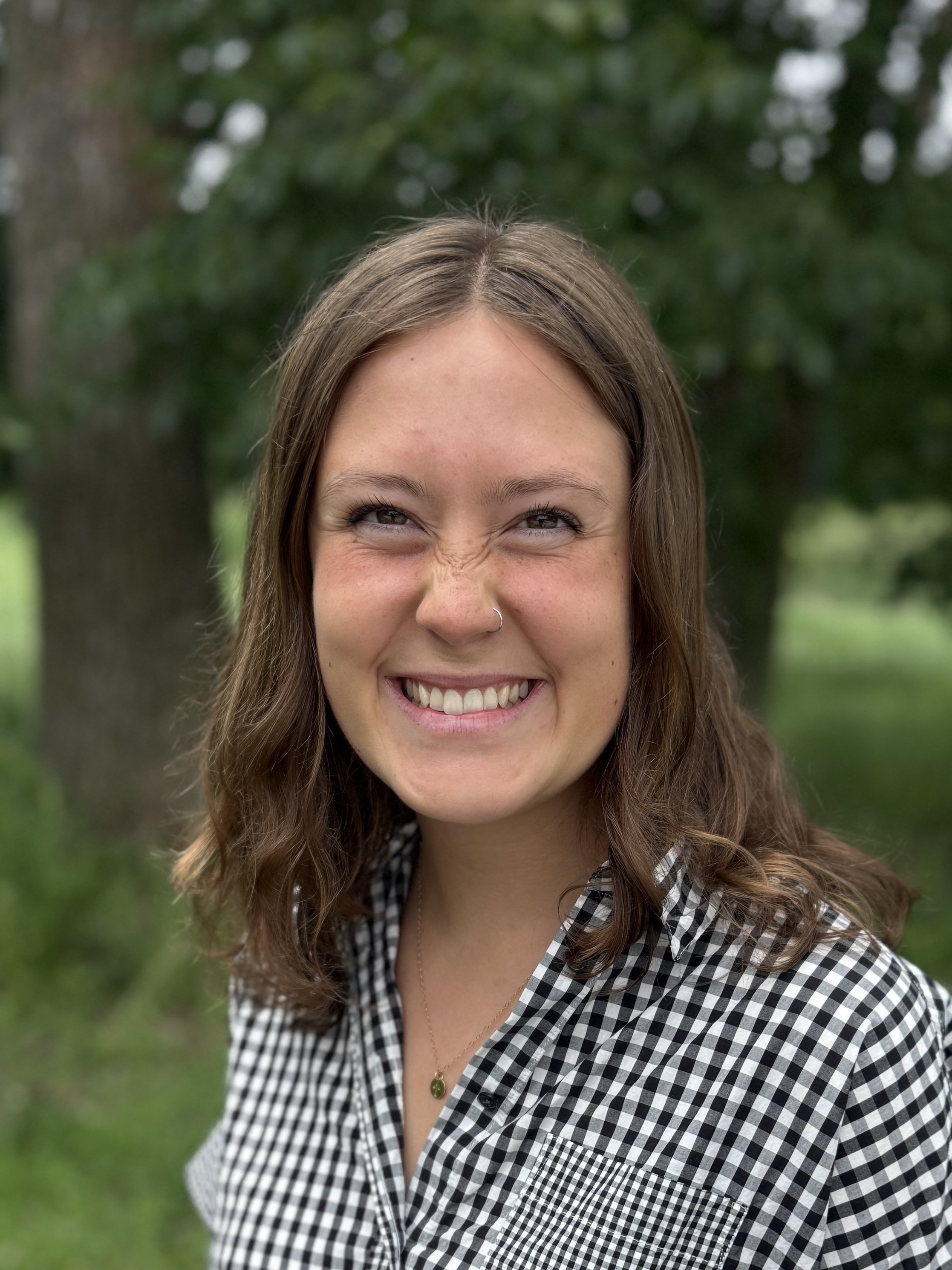A woman with curly dark hair smiling outdoors, wearing a blue striped short-sleeve top, with a blurred green background.