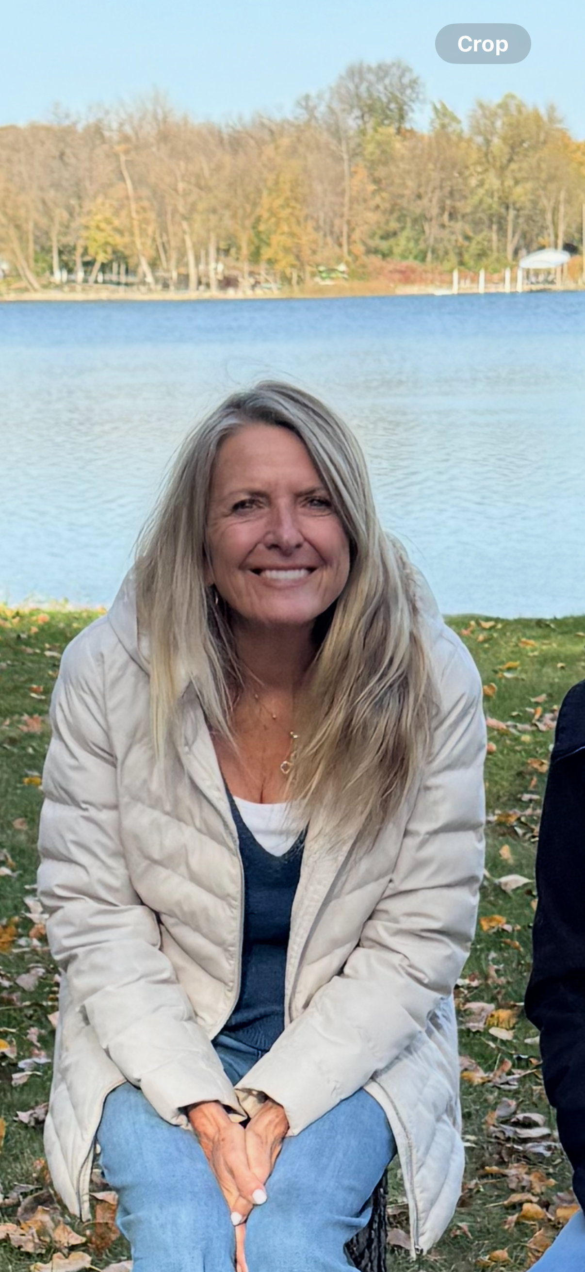 Portrait of a smiling man with short dark hair and a beard, wearing a light grey blazer and black shirt, standing outdoors with blurred green trees in the background.