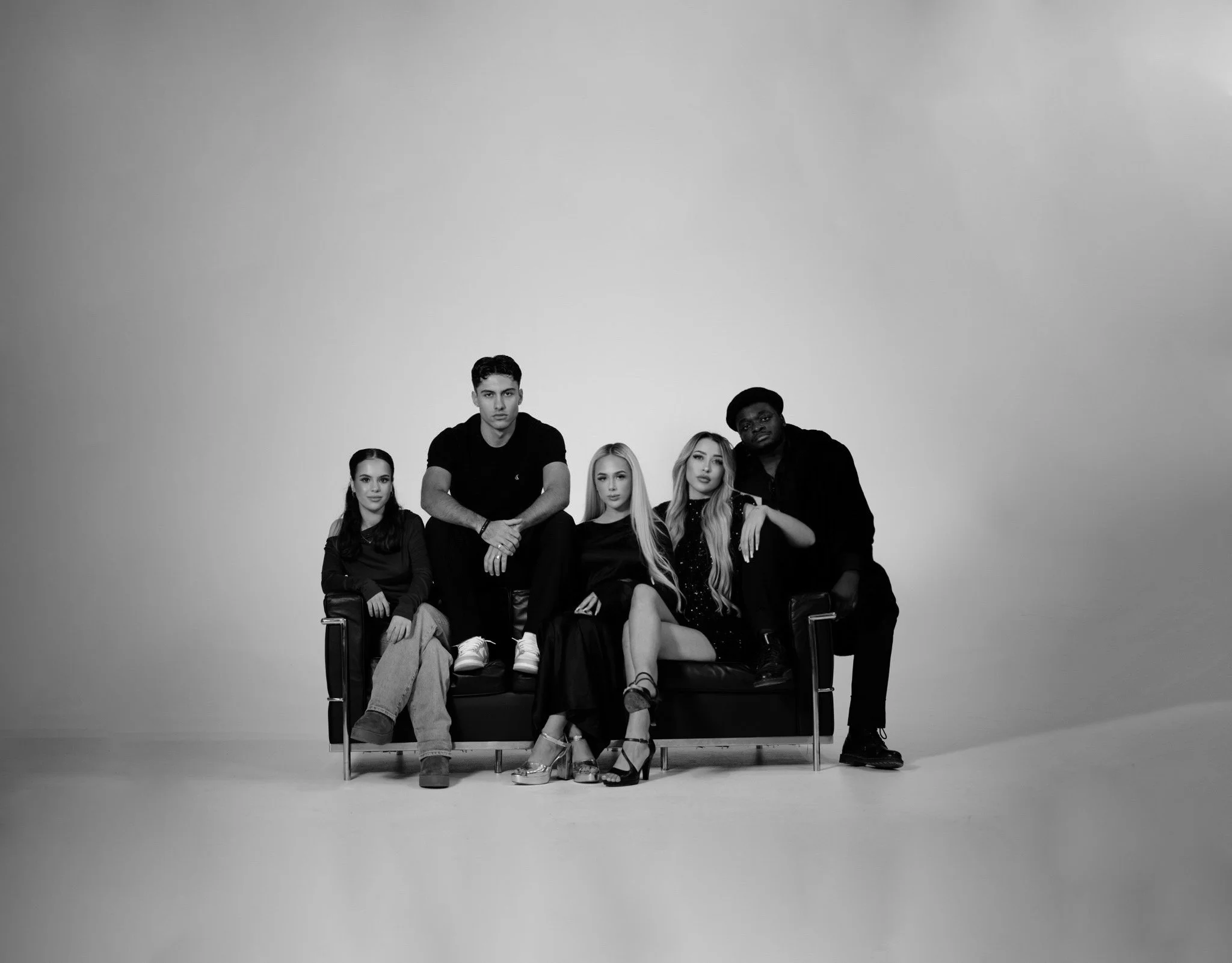 Fünf junge Menschen sitzen und stehen auf einer schwarzen Couch vor einem weißen Hintergrund, alle in schwarzen Kleidern und modernem Stil, schwarz-weiß Fotografie.