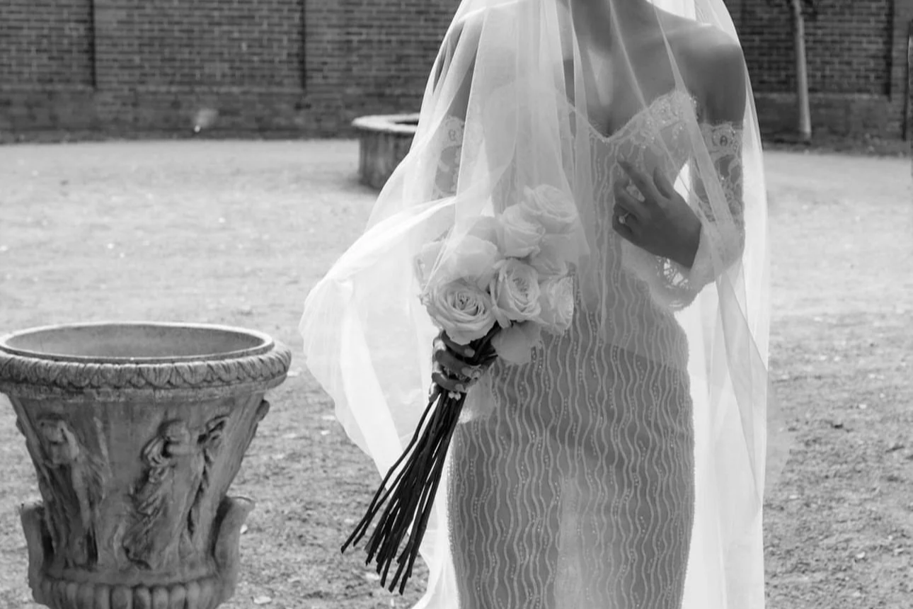 Black and white photo of a bride holding a bouquet of roses, partially covered by a veil, outdoors with a brick wall background.