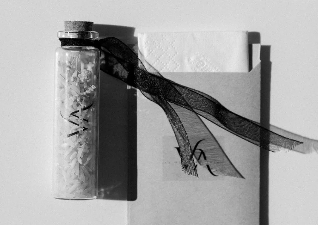 A glass jar filled with small paper pieces, sealed with a cork, resting on a surface. Behind it are a box of tissues and a ribbon tied around the jar, with the box partially obscured.