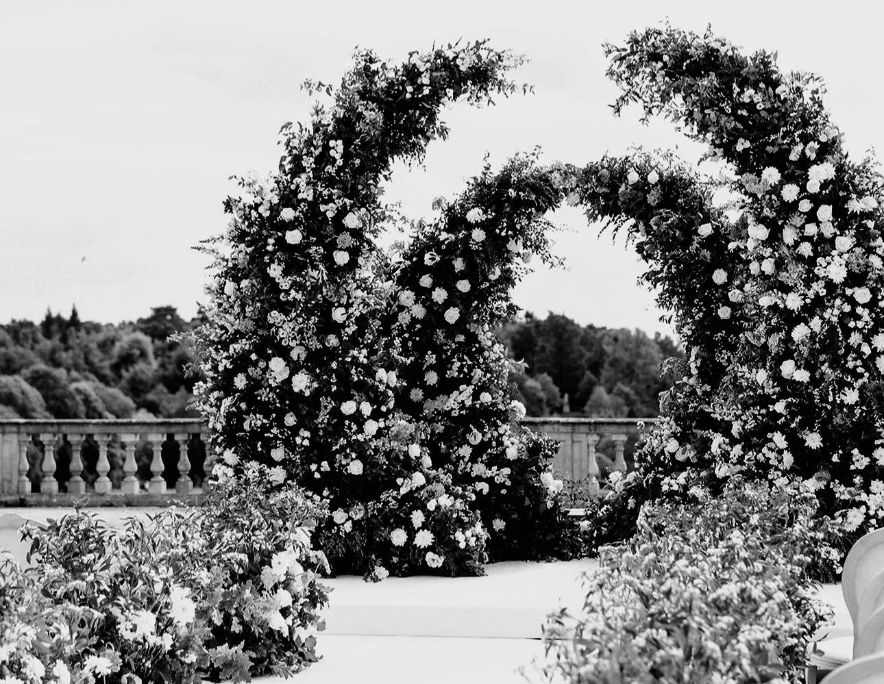 Decorative floral arch made of flowers and greenery, set outdoors with a stone railing and trees in the background.