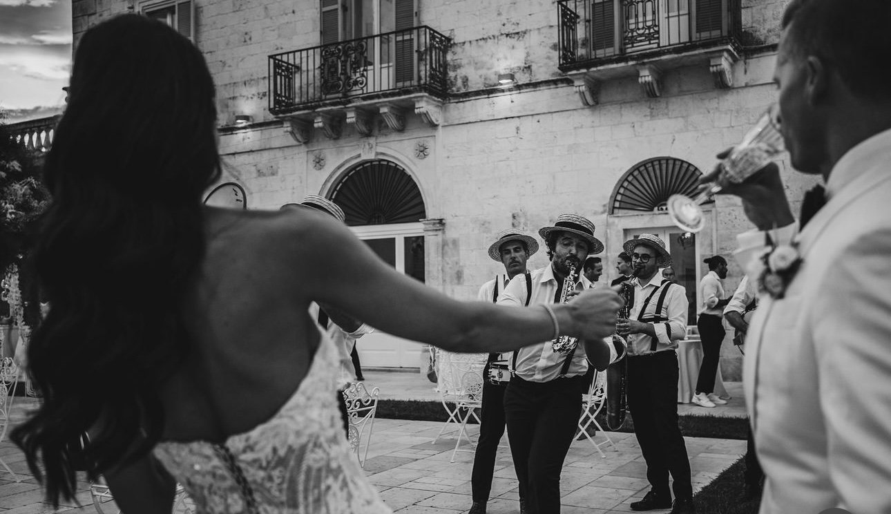 People dancing and playing music at an outdoor wedding reception in the evening, with a woman in a dress and a man in a tuxedo raising glasses.