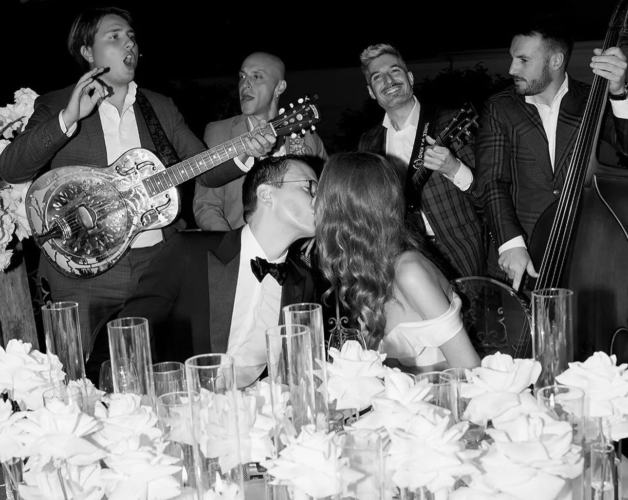 A black and white photo of a wedding celebration with a bride and groom kissing at a table surrounded by friends playing musical instruments and singing, with candles and flowers on the table.