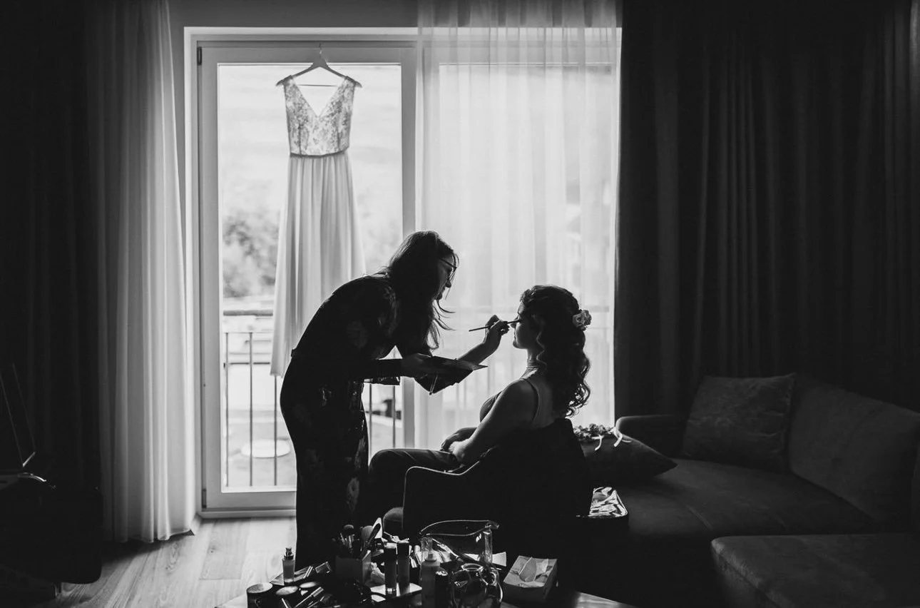 A woman getting her makeup done indoors, with a wedding dress hanging on a hanger in the window behind her.