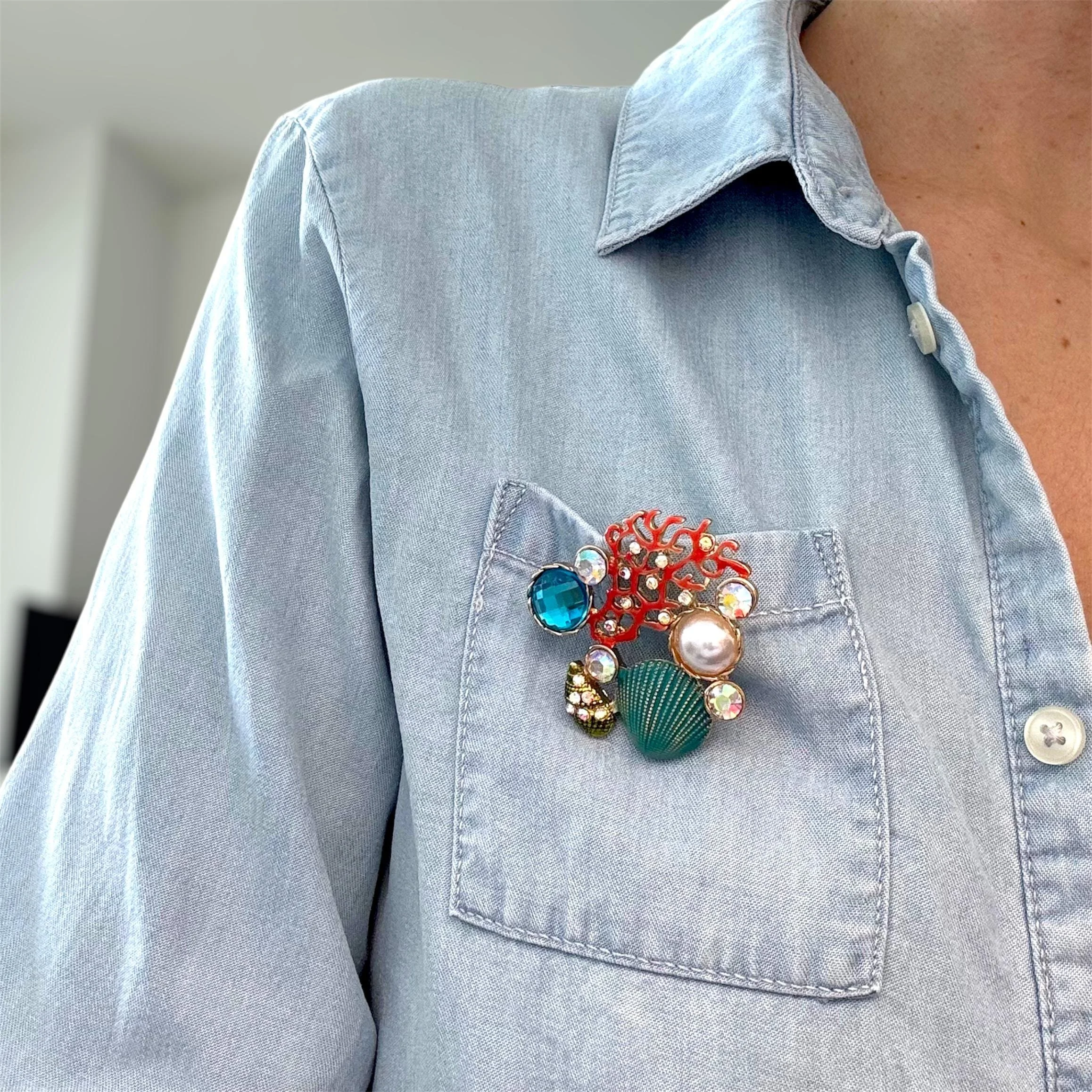 Brooch mit Muschel, Perlen, Edelsteinen und roten Mustern an einer Jeansjacke befestigt.