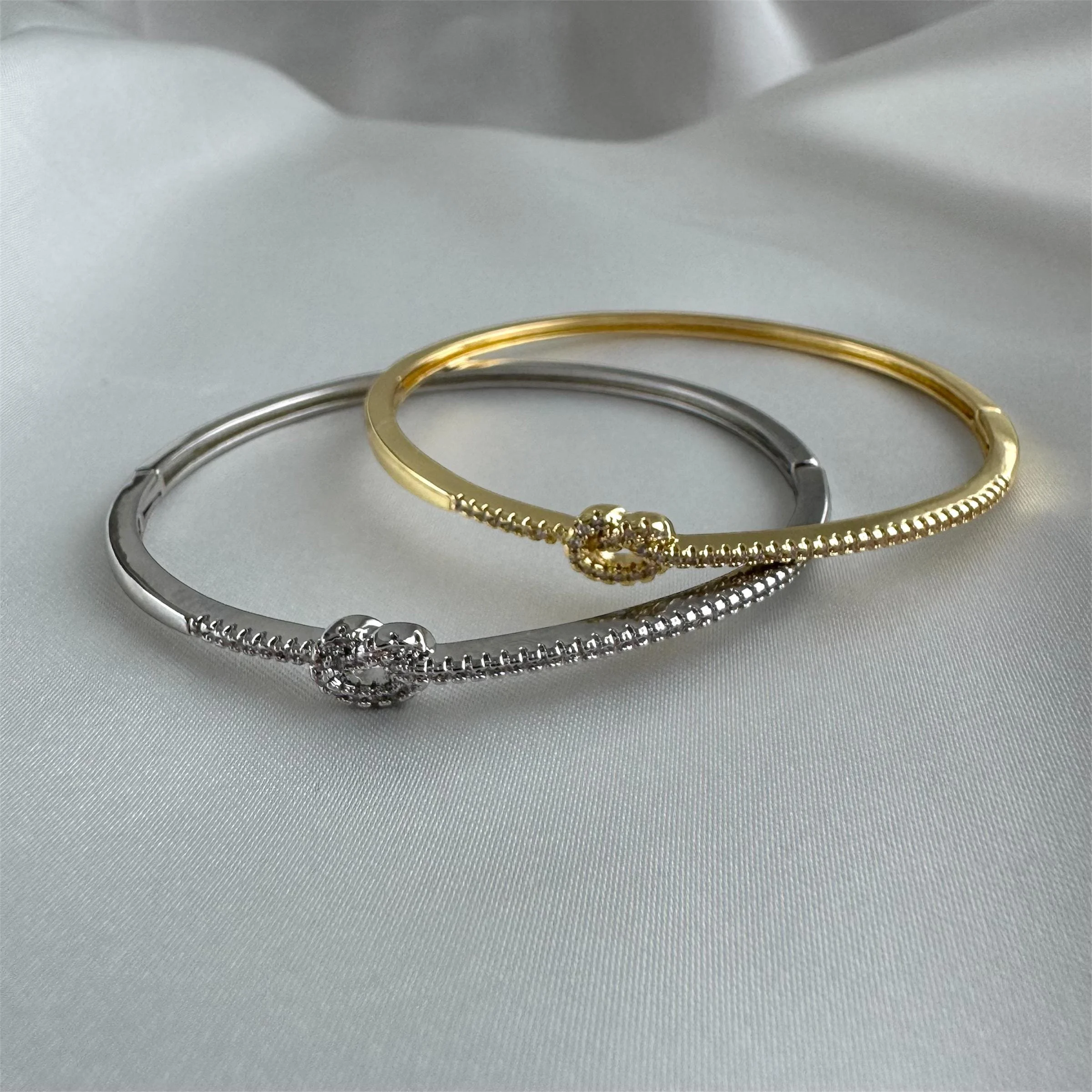 Zwei schlichte Armbänder, eines in Silber und das andere in Gold, mit kleinen, kugelähnlichen Verzierungen, liegen auf weißem Satin.