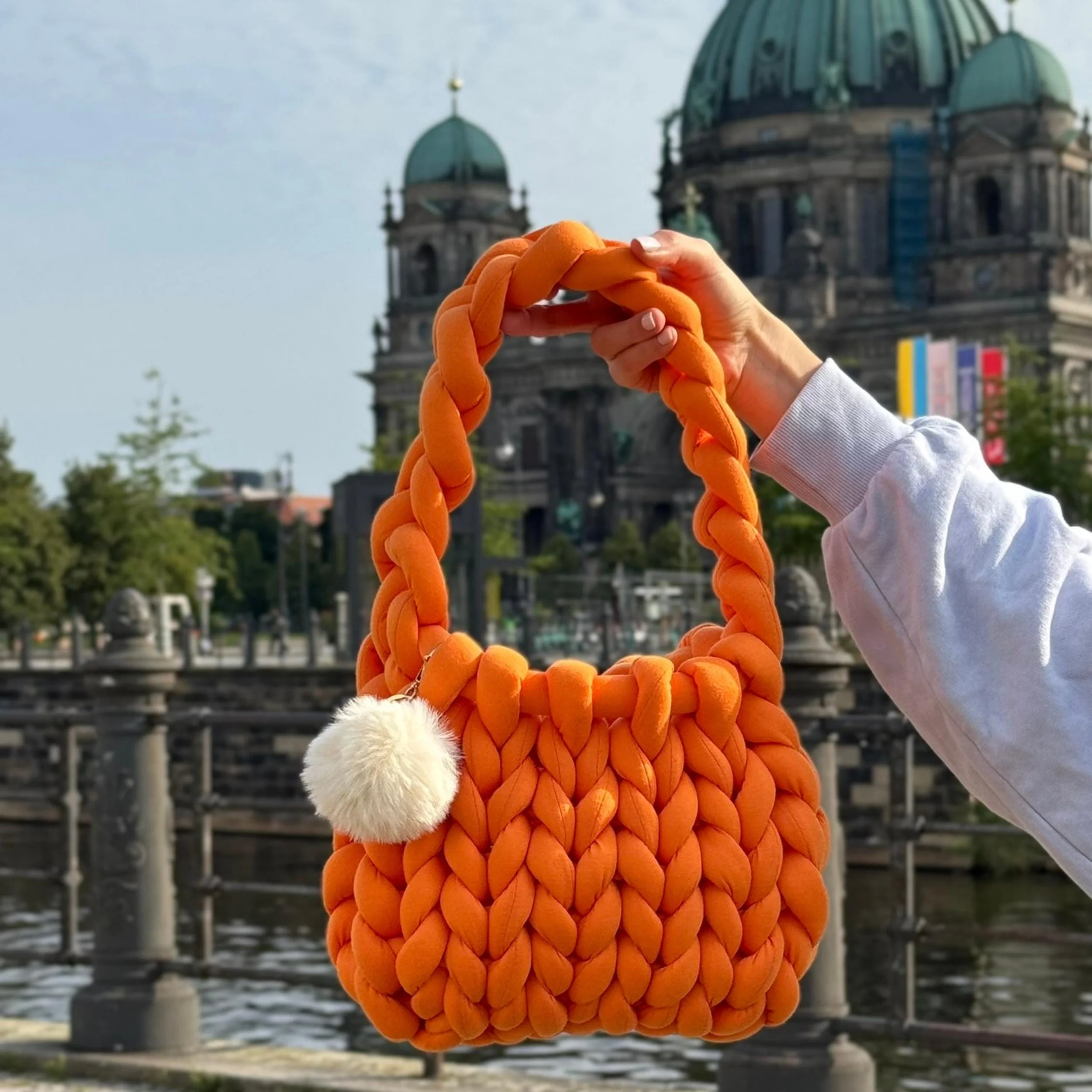 Eine orange, geflochtene Handtasche mit einem weißen Pompon-Anhänger, gehalten von einer Person vor einer historischen Gebäudeansicht in Berlin.