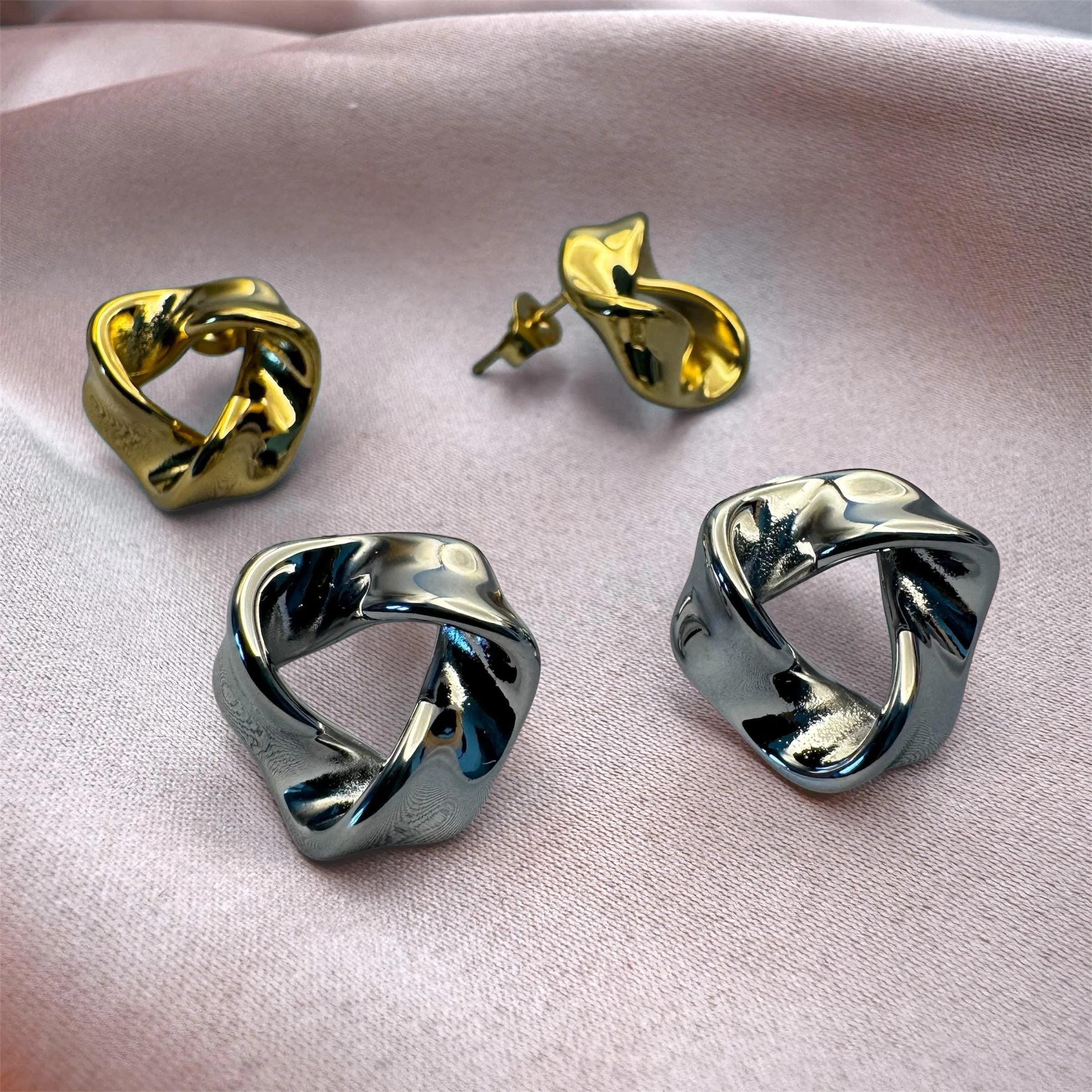 Zwei silberne und zwei goldene, modern geformte Ohrringe auf einem pinken Satinuntergrund.