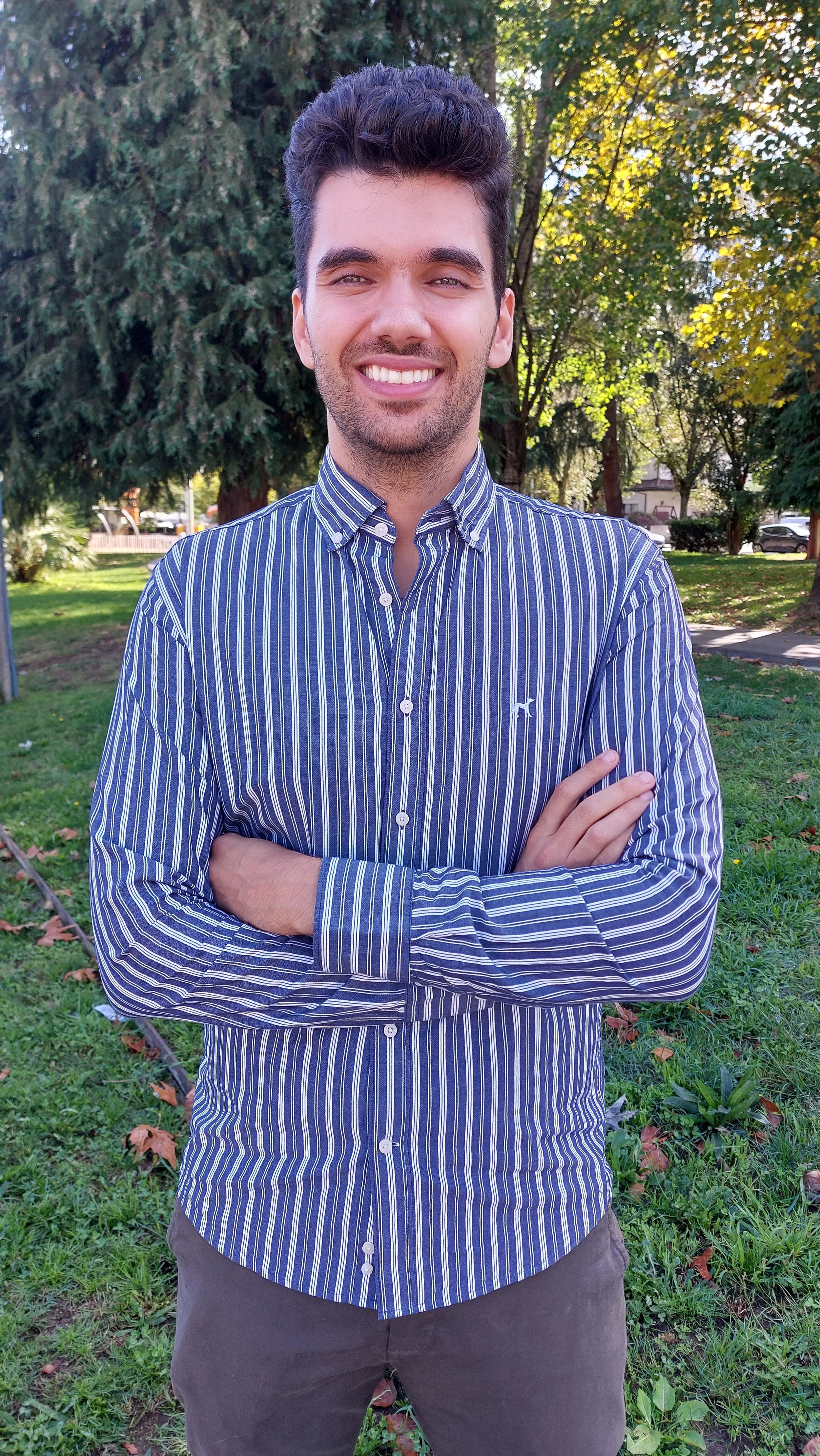 A young man with dark hair and a beard, smiling with his arms crossed, wearing a blue and white striped button-up shirt, standing outdoors in a park with green grass, trees, and sunlight.