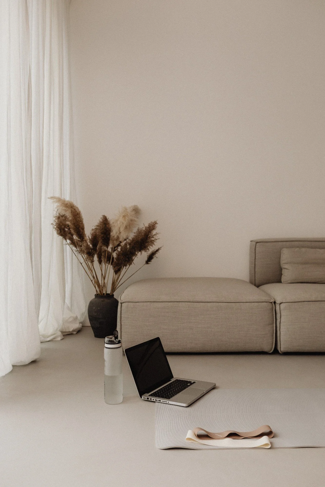 Minimalist living room with beige sofa, vases with dried pampas grass, a laptop, water bottle, yoga mat, and a towel on a neutral-colored carpet.