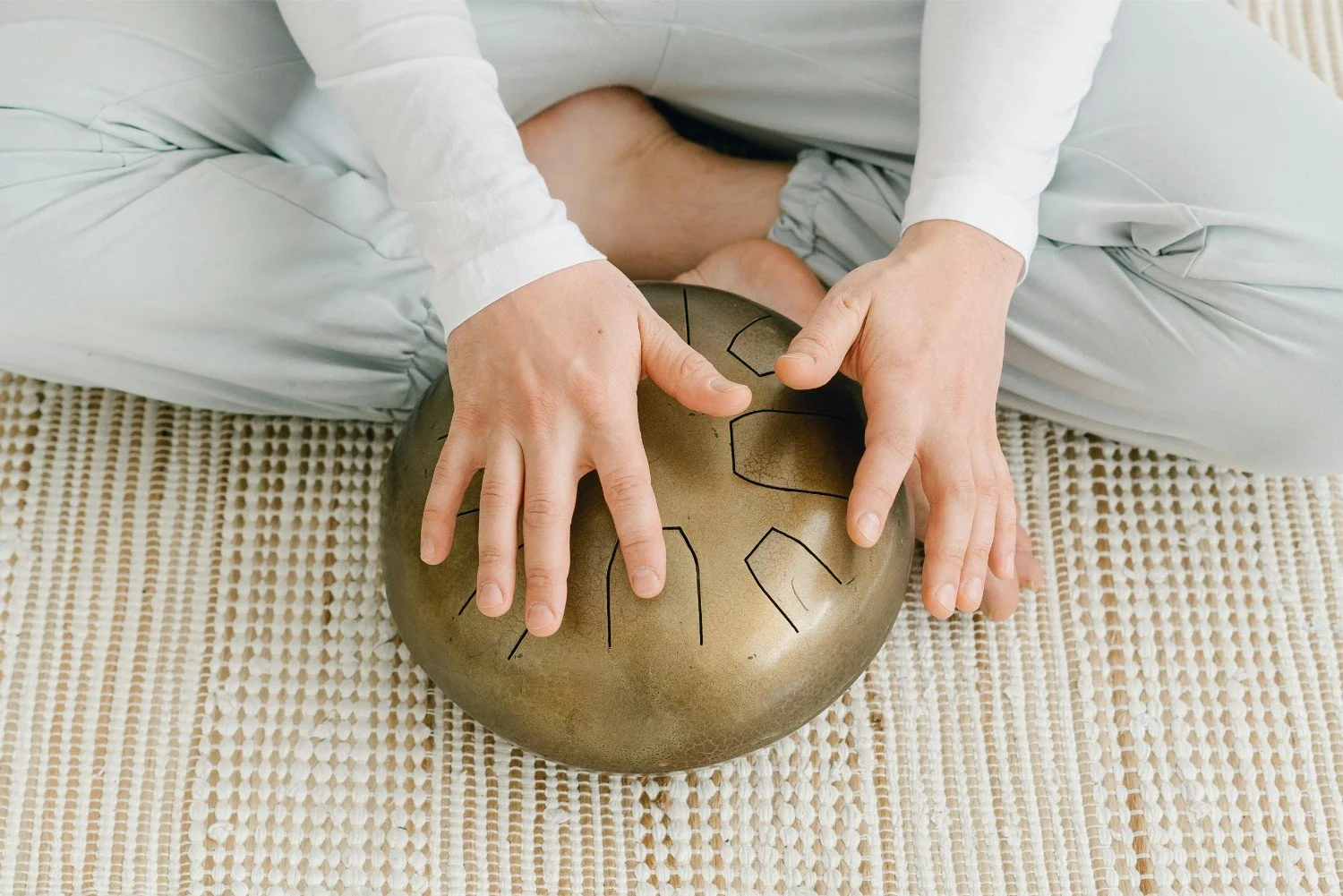 Person playing a tongue drum with their hands while sitting on a woven mat.