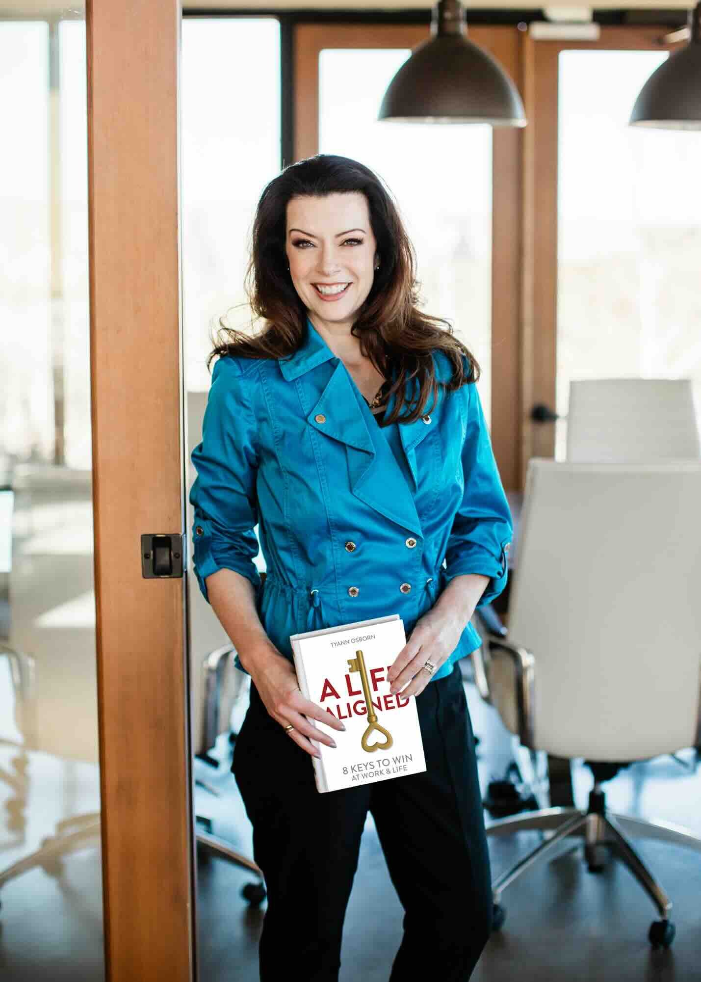 A woman with long dark hair, wearing a bright blue jacket, smiling and holding a book titled 'ALIGNED' in an office or conference room setting.