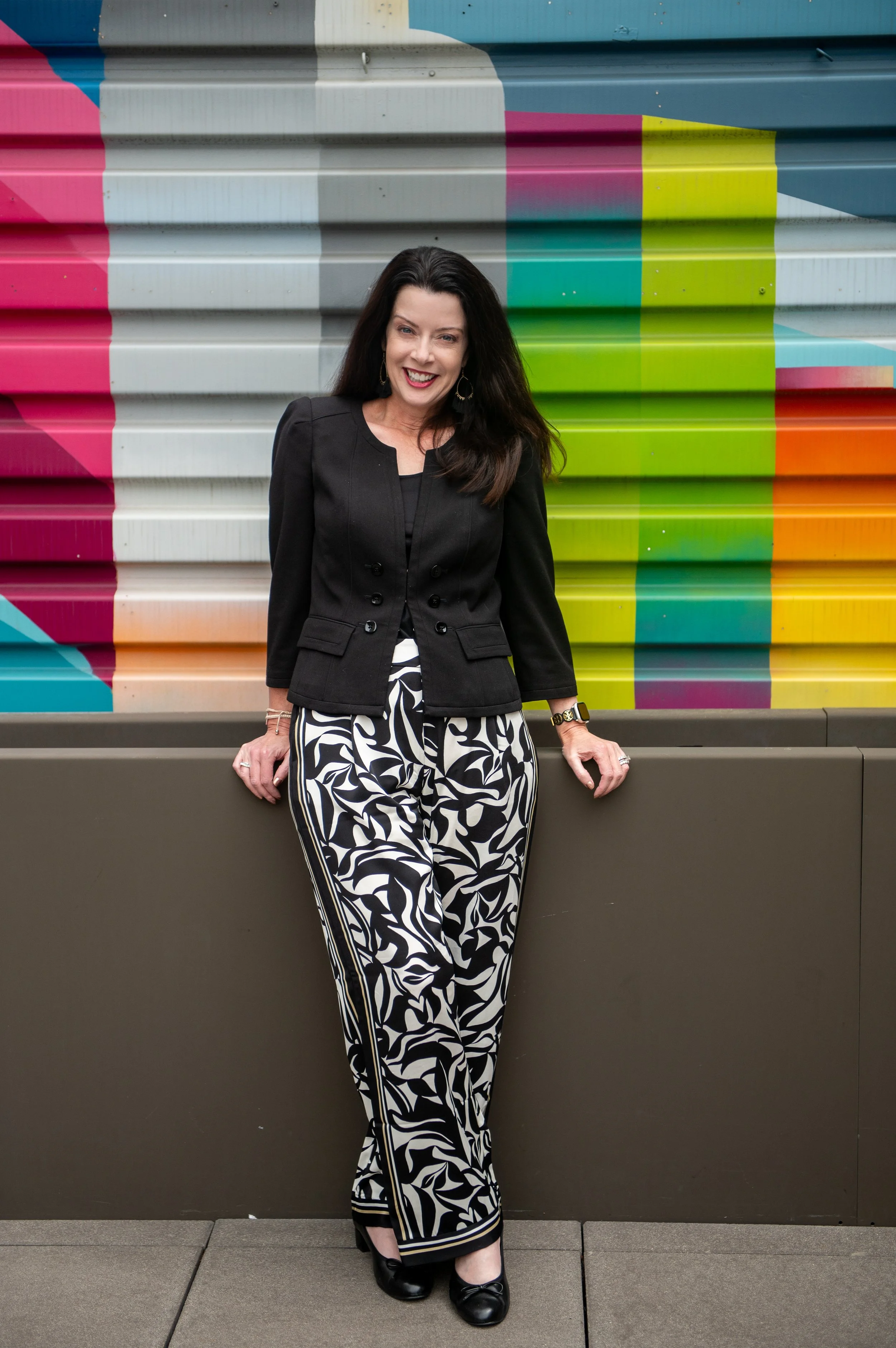 A woman standing against a colorful mural wall, wearing a black blazer, patterned black and white pants, and black shoes.