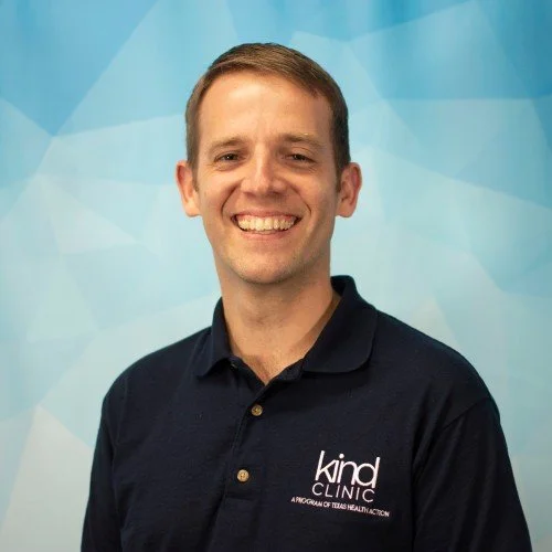 A smiling man with short brown hair wearing a black polo shirt with a 'Kind Clinic' logo standing in front of a blue abstract background.