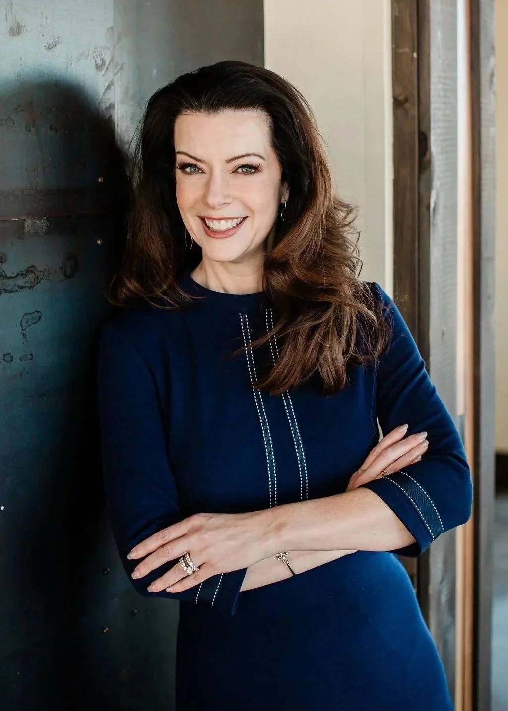 A woman with long, wavy brown hair, smiling, wearing a navy blue dress with white stitching, standing cross-armed near a dark wall with a concrete and wood background.