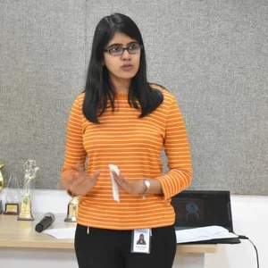 Young woman with glasses standing in front of a gray wall, holding a notepad, wearing an orange striped sweater and black pants.