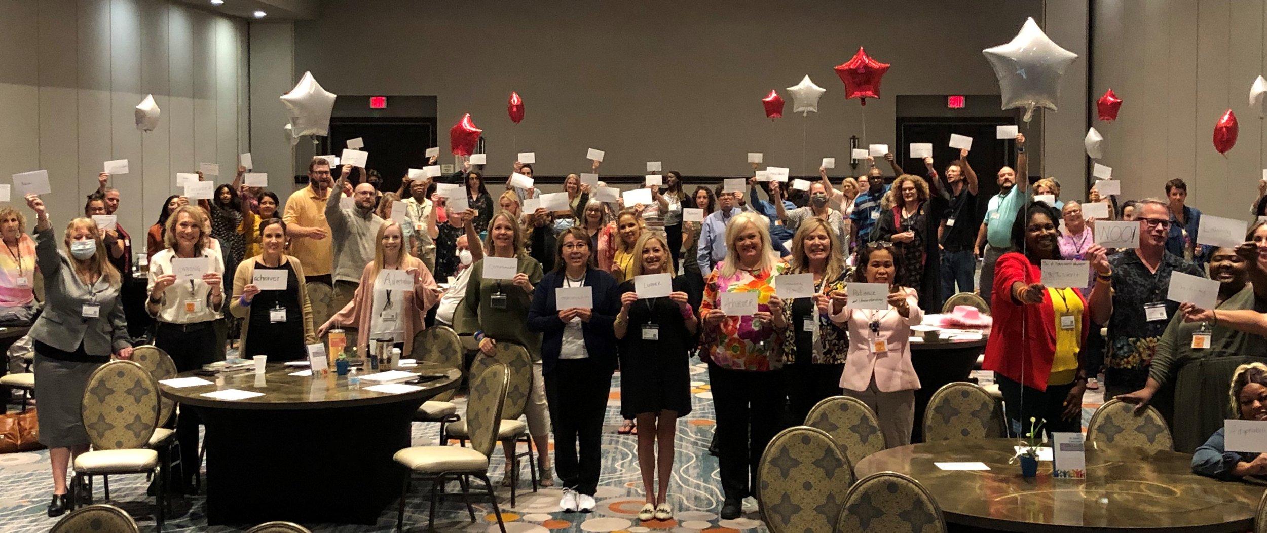 A large group of people in a conference room holding white cards, with red and white star-shaped balloons floating overhead.