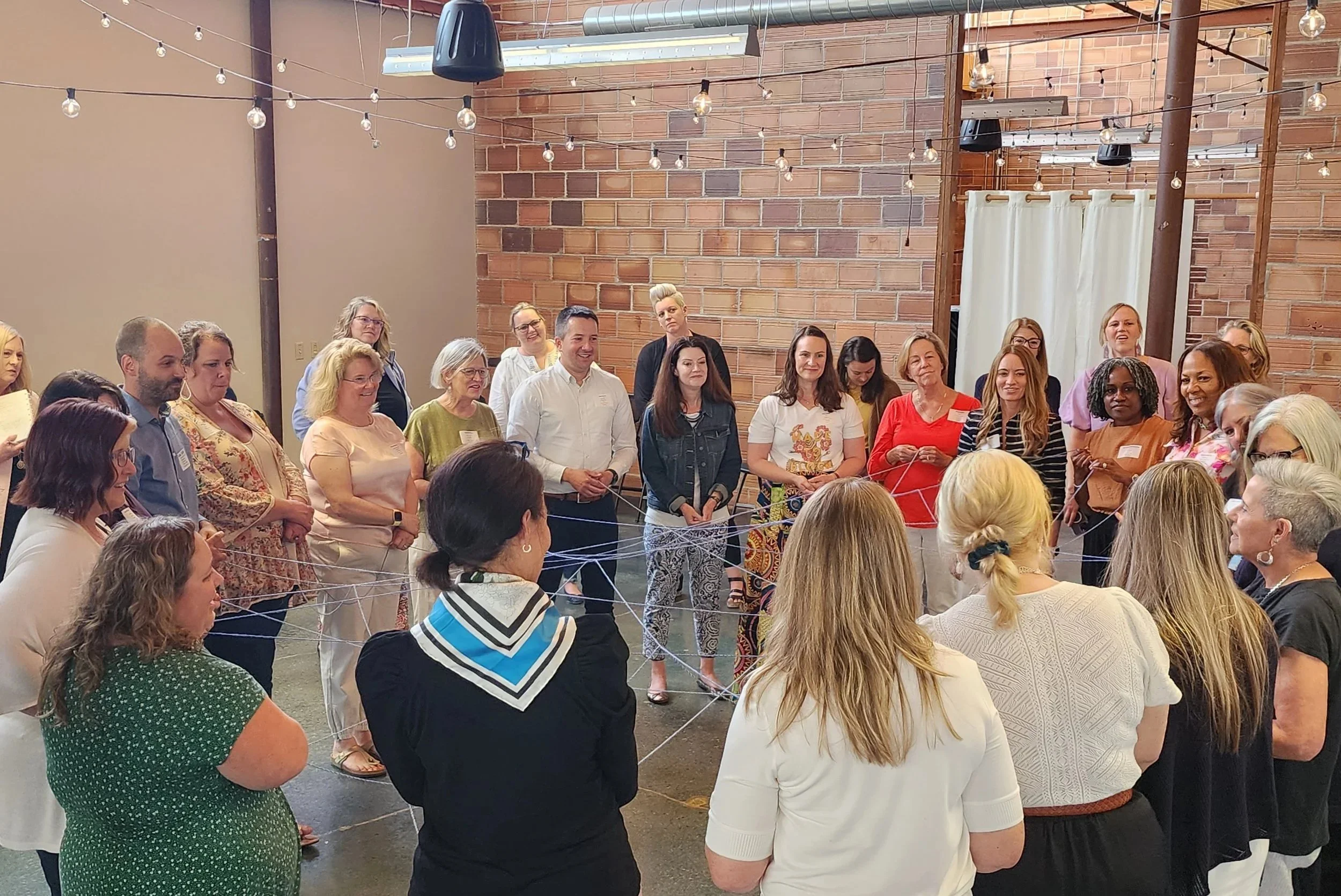 Group of adults participating in a team-building activity, standing in a circle with strings connecting them, in an indoor space with brick walls, string lights, and a curtain in the background.