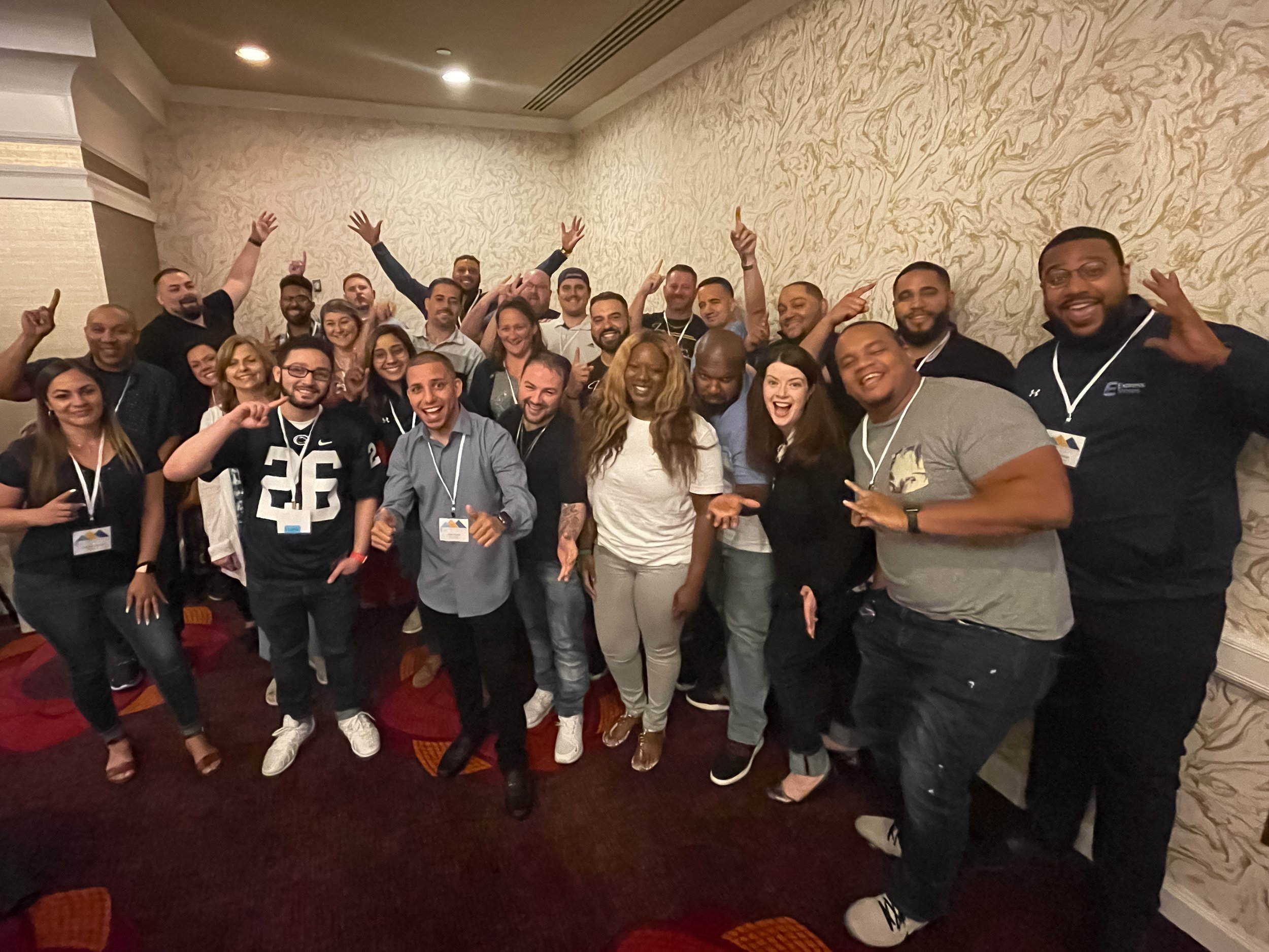 Group of smiling people posing together in a room with beige patterned walls. Some are wearing lanyards and casual clothing.