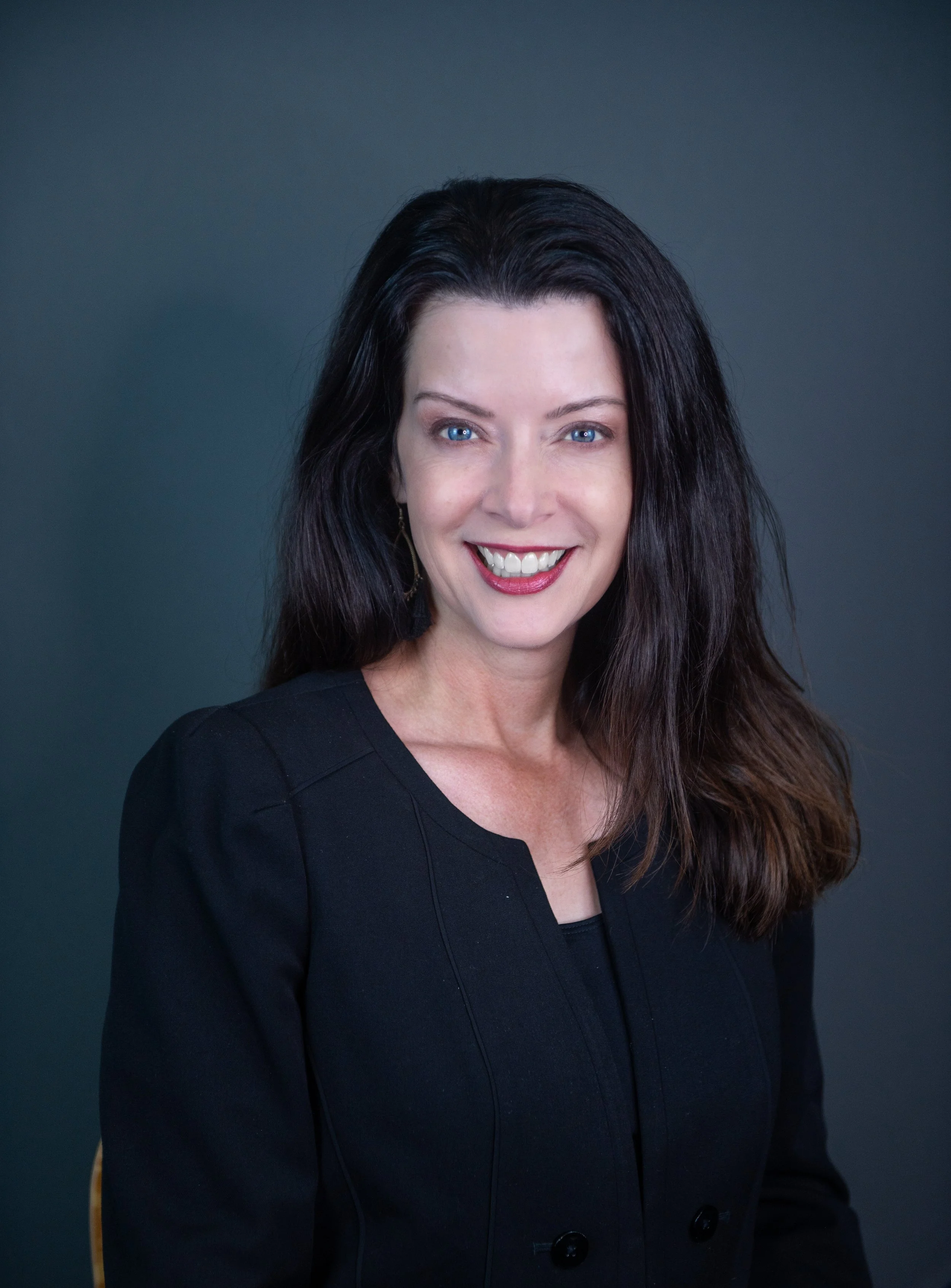 Portrait of a woman with long dark hair wearing a black blazer, smiling against a dark background.