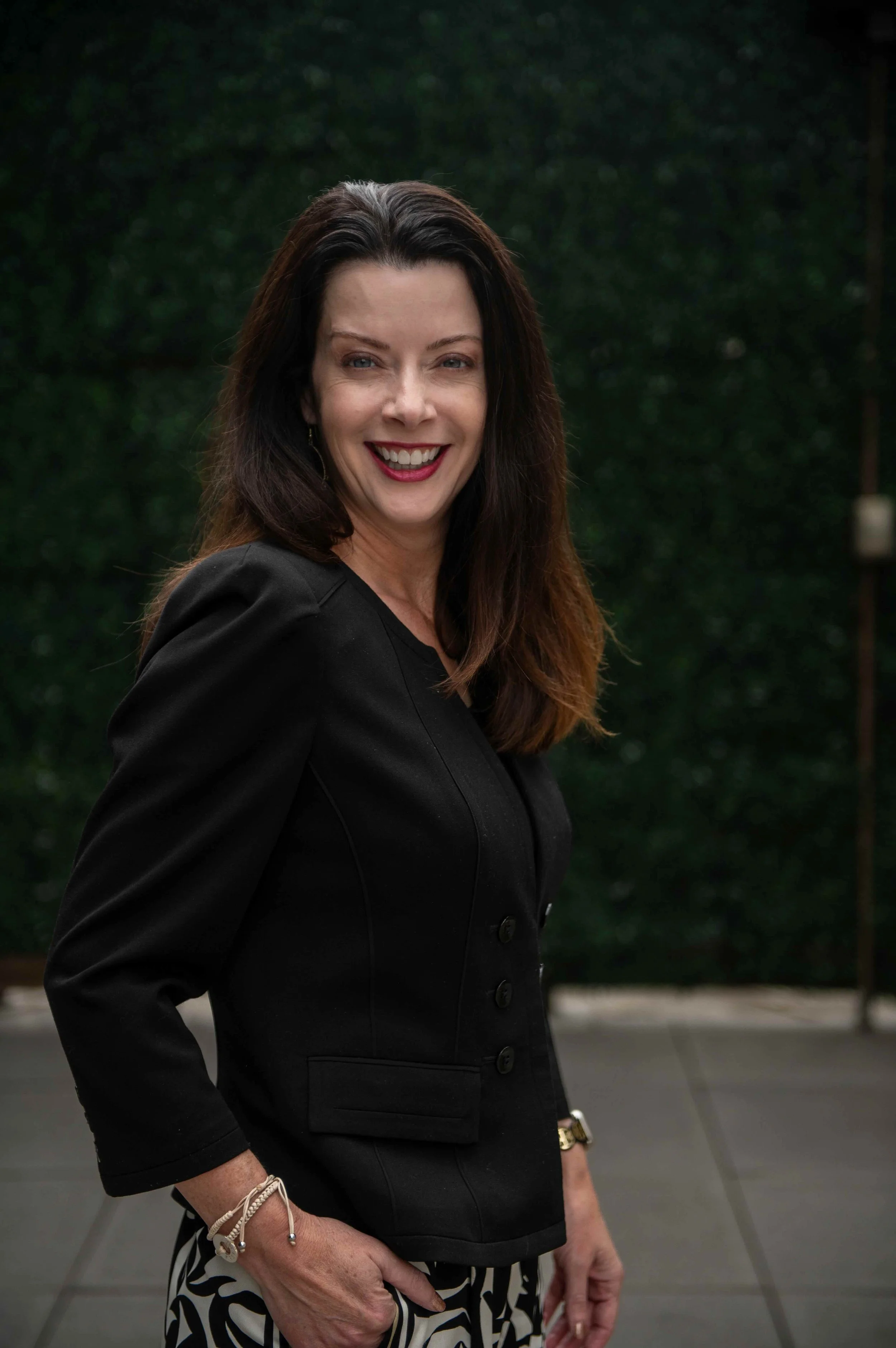 A woman with dark hair smiling, wearing a black blazer and patterned pants, standing outdoors with a blurred green hedge background.
